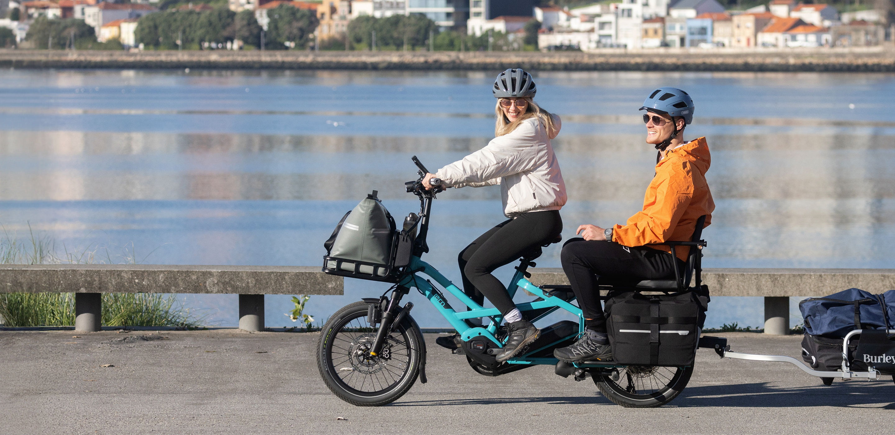 A girl comfortably riding a Tern GSD with a guy on the passenger seat, and a trailer to haul cargo.