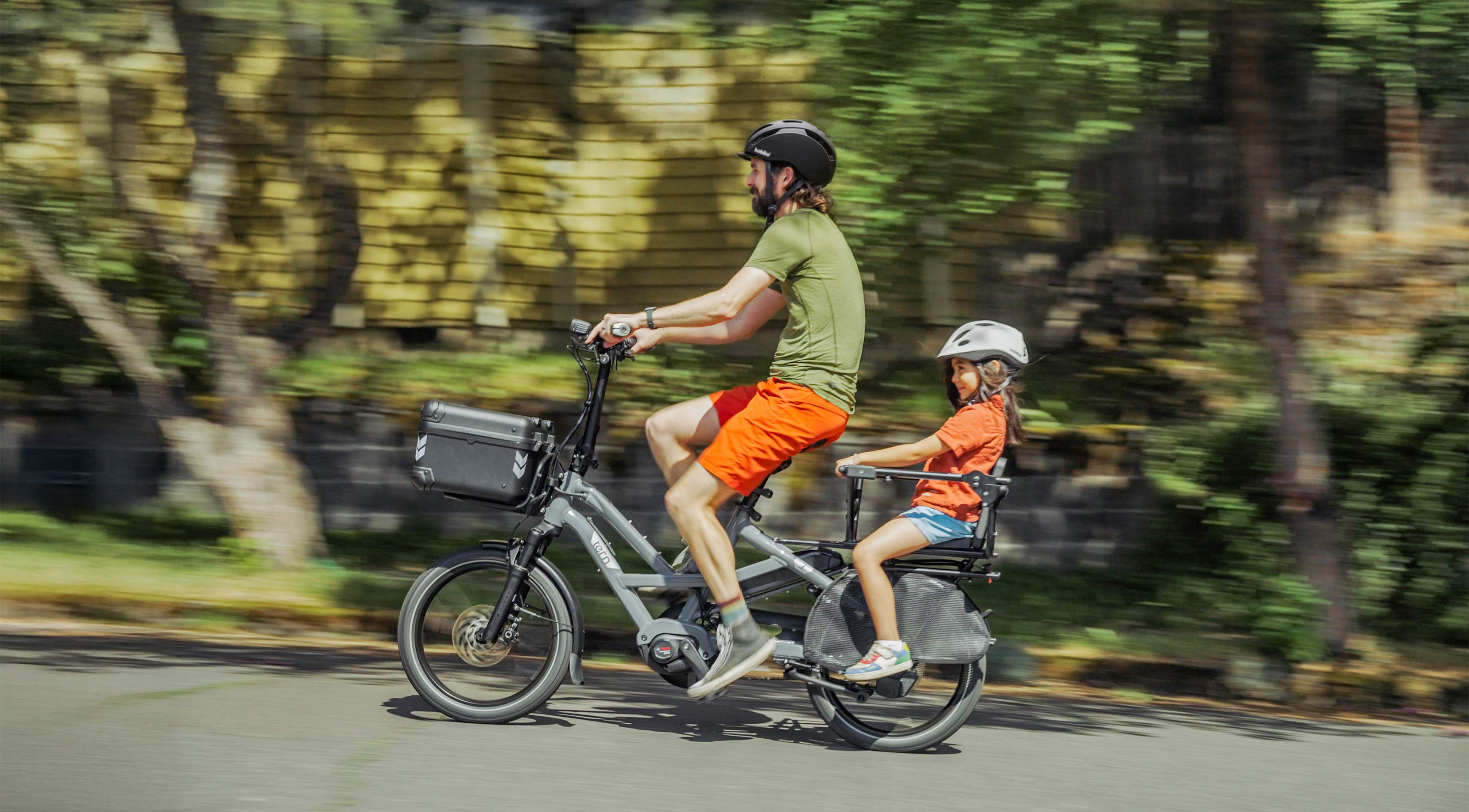 A man rides a Tern HSD with an adult passenger on the rear seat and a dog in the front pet carrier.